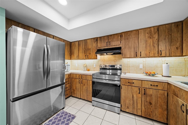 kitchen with light tile patterned floors, stainless steel appliances, under cabinet range hood, brown cabinets, and backsplash