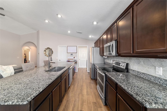 kitchen with dark stone countertops, an island with sink, a sink, vaulted ceiling, and appliances with stainless steel finishes