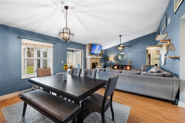 dining space with baseboards, lofted ceiling, a brick fireplace, ceiling fan with notable chandelier, and light wood-type flooring