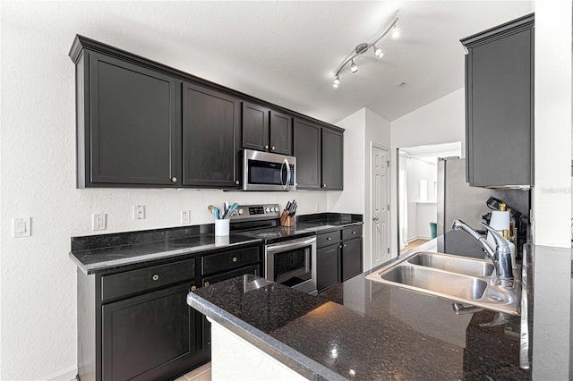 kitchen with track lighting, lofted ceiling, dark stone countertops, appliances with stainless steel finishes, and a sink