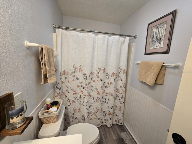 bathroom with a shower with shower curtain, wood finished floors, a wainscoted wall, a textured ceiling, and toilet