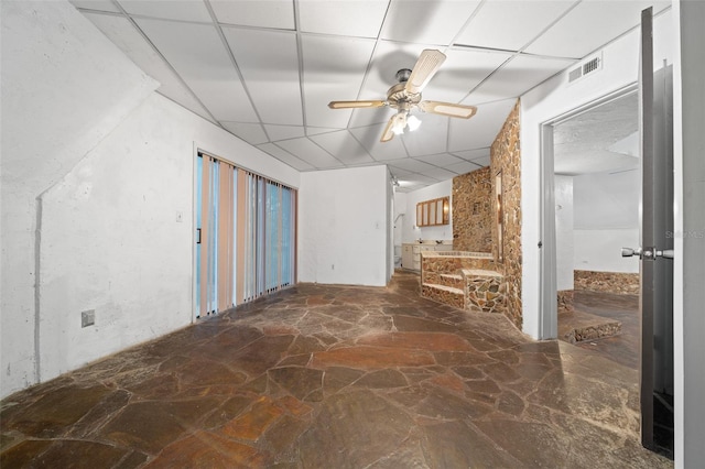 empty room featuring a ceiling fan, visible vents, a drop ceiling, and stone floors