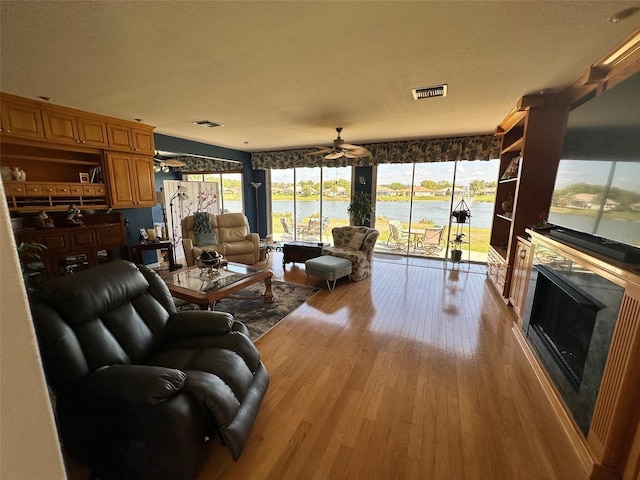 living area with wood finished floors, visible vents, ceiling fan, a water view, and a glass covered fireplace