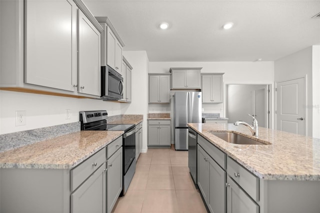 kitchen featuring an island with sink, stainless steel appliances, gray cabinets, a sink, and light stone countertops