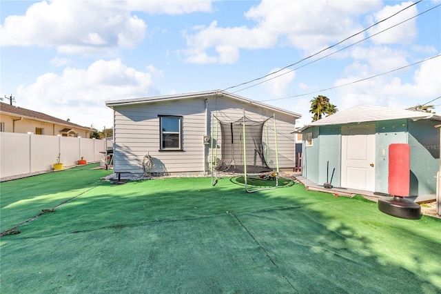 back of property featuring a trampoline, an outdoor structure, fence, and a lawn