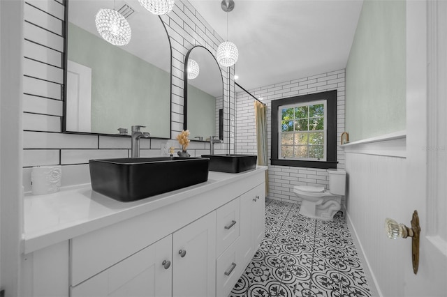 full bathroom featuring toilet, vanity, tile walls, a chandelier, and a wainscoted wall