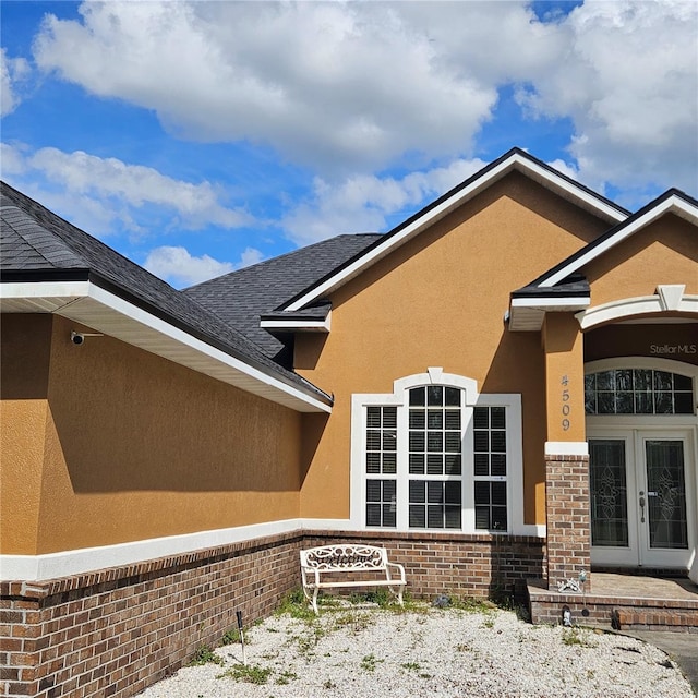 doorway to property featuring a shingled roof, brick siding, stucco siding, and french doors