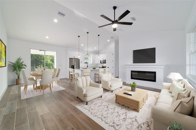 living area featuring vaulted ceiling, ceiling fan, a glass covered fireplace, baseboards, and wood finished floors