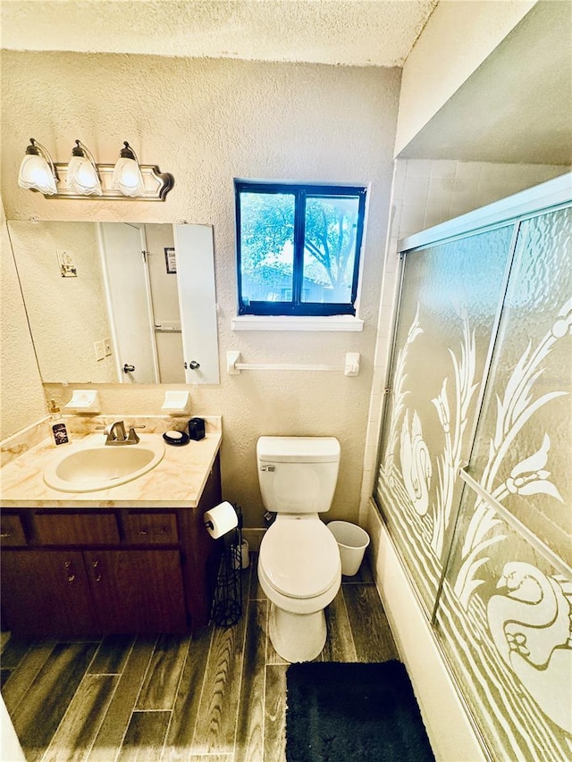 full bath featuring toilet, a textured wall, vanity, wood finished floors, and a shower with door