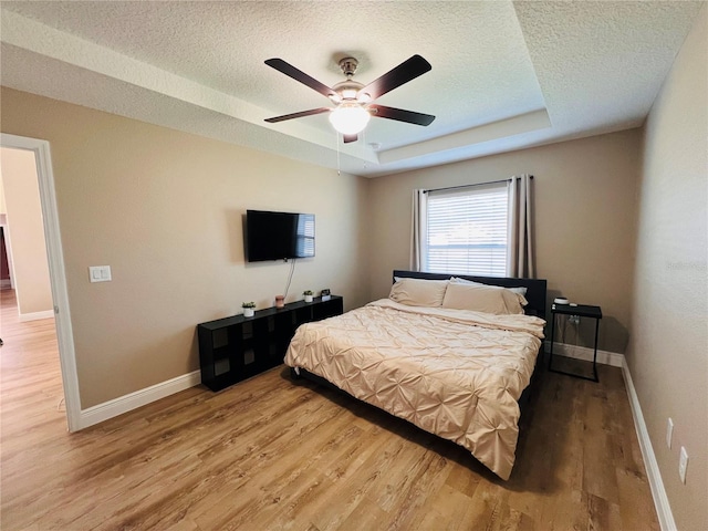 bedroom featuring a raised ceiling, a ceiling fan, light wood-type flooring, baseboards, and a textured ceiling