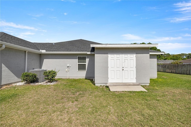 rear view of house with fence, a shingled roof, central AC, stucco siding, and a lawn