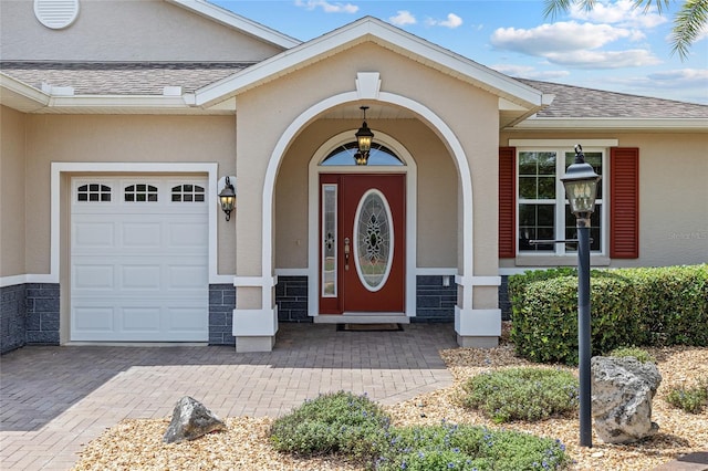 view of exterior entry with stucco siding, roof with shingles, decorative driveway, and a garage