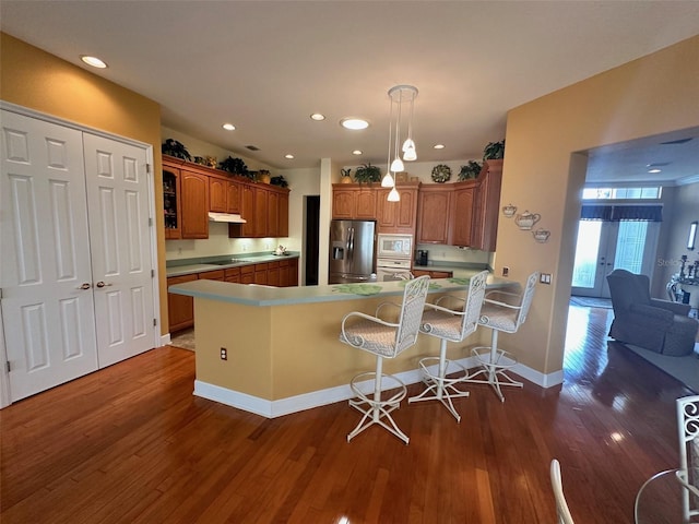 kitchen with dark wood finished floors, under cabinet range hood, white appliances, a peninsula, and brown cabinetry