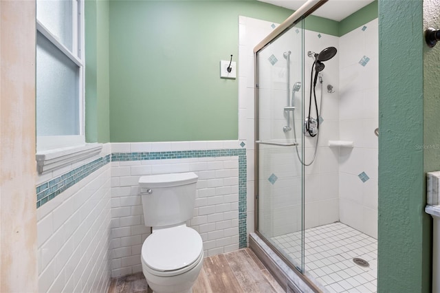 bathroom with wainscoting, toilet, tile walls, a shower stall, and wood finished floors