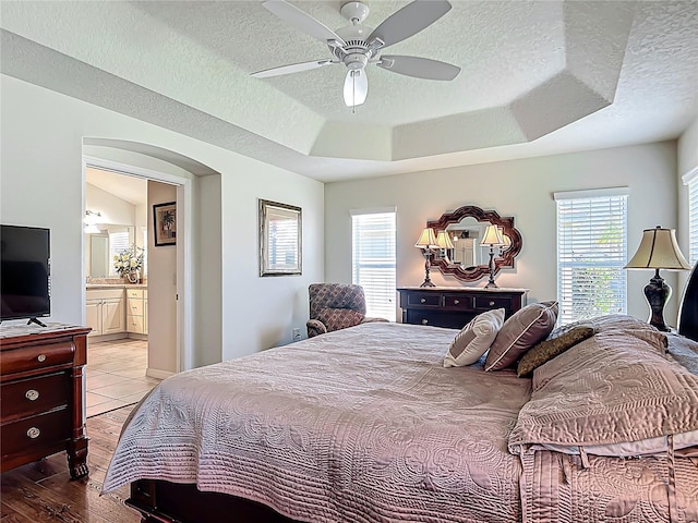 bedroom with a raised ceiling, a textured ceiling, a ceiling fan, light wood-style flooring, and ensuite bath