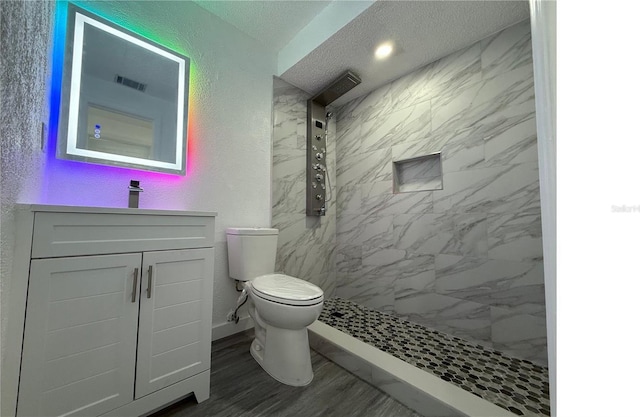 full bathroom with vanity, toilet, a tile shower, visible vents, and a textured wall