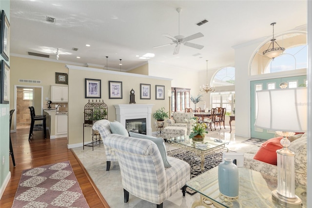 living room featuring crown molding, visible vents, a glass covered fireplace, wood finished floors, and ceiling fan with notable chandelier