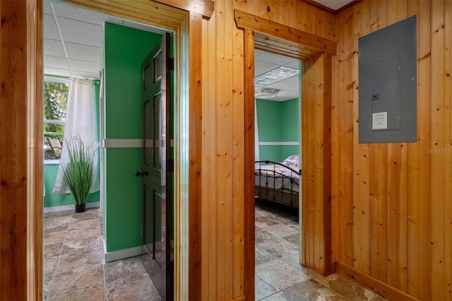 hallway with wood walls, stone finish flooring, electric panel, baseboards, and a paneled ceiling