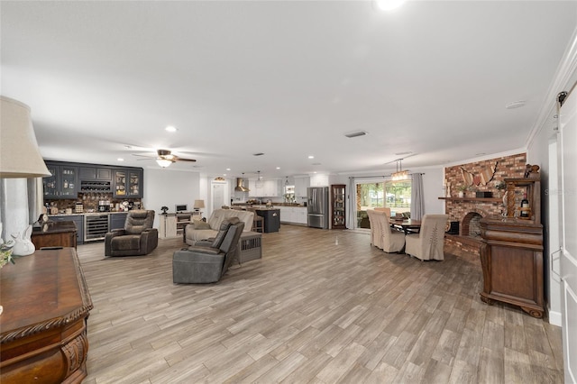 living area featuring a ceiling fan, a brick fireplace, light wood-style floors, ornamental molding, and recessed lighting