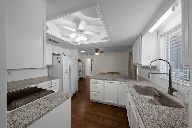 kitchen featuring a sink, white refrigerator with ice dispenser, a ceiling fan, a peninsula, and a raised ceiling