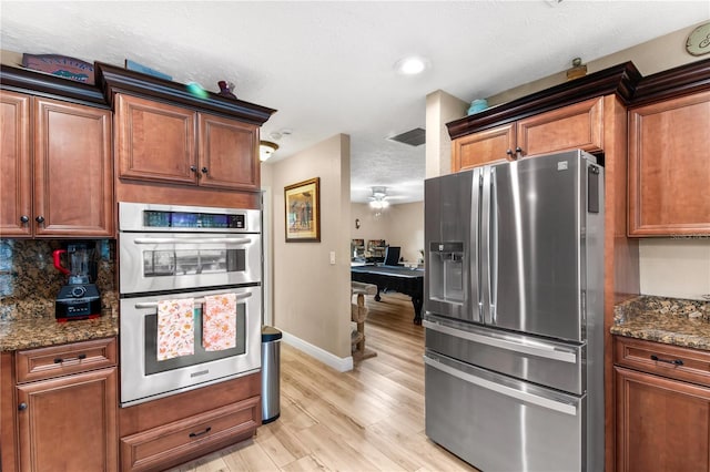 kitchen featuring appliances with stainless steel finishes, light wood-style floors, brown cabinetry, dark stone counters, and baseboards