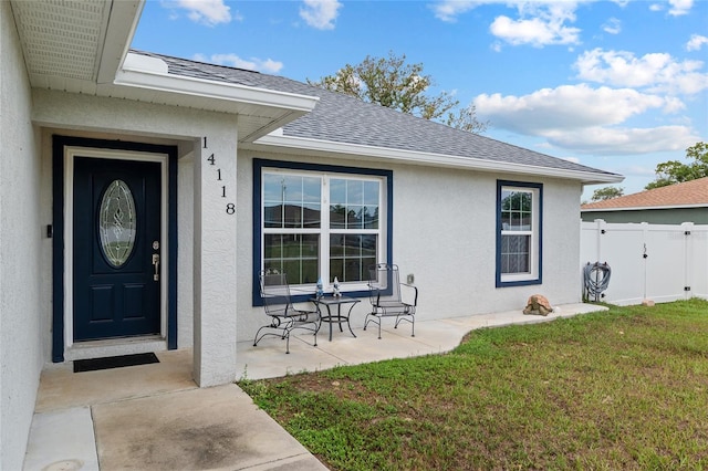 property entrance featuring fence, stucco siding, a yard, and a shingled roof