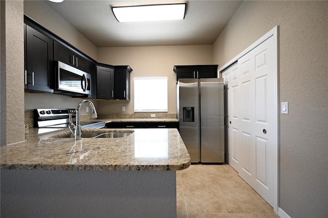 kitchen with light stone countertops, dark cabinetry, a sink, a peninsula, and stainless steel appliances