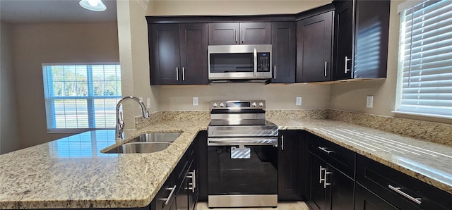 kitchen with a peninsula, dark brown cabinets, a sink, appliances with stainless steel finishes, and light stone counters