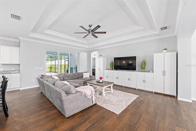 living room featuring a tray ceiling, a ceiling fan, dark wood-style flooring, crown molding, and baseboards