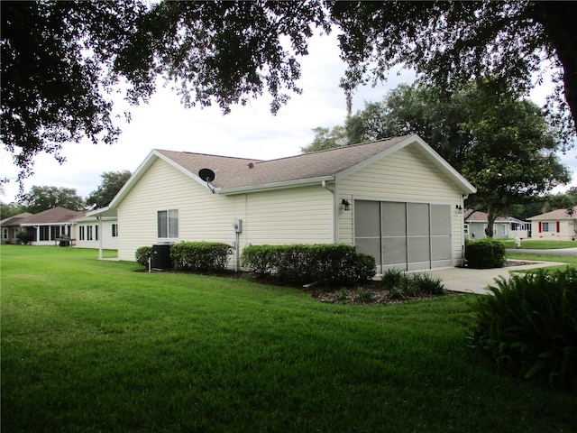 view of side of property featuring an attached garage, a lawn, central AC unit, roof with shingles, and driveway