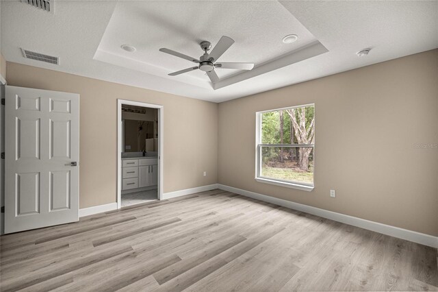unfurnished bedroom featuring a tray ceiling, light wood finished floors, baseboards, and ensuite bathroom