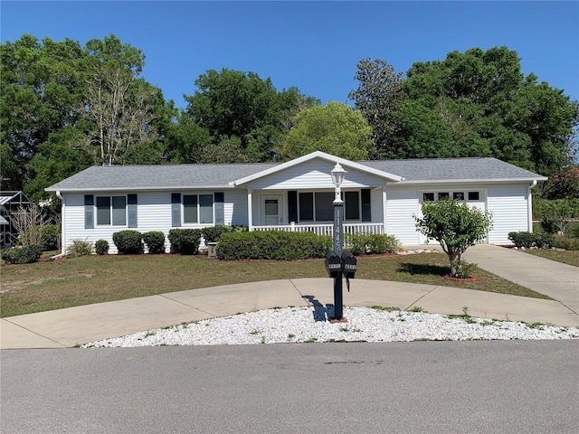 ranch-style home featuring covered porch, a front lawn, driveway, view of wooded area, and roof with shingles