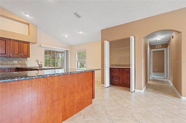 kitchen with dark stone countertops, arched walkways, decorative backsplash, lofted ceiling, and brown cabinets