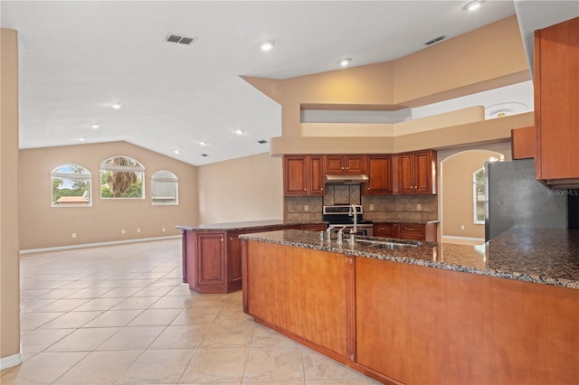 kitchen with a peninsula, appliances with stainless steel finishes, under cabinet range hood, backsplash, and dark stone countertops