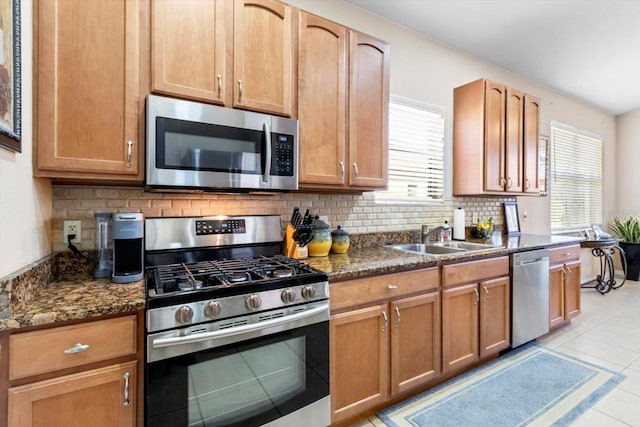 kitchen with appliances with stainless steel finishes, a sink, backsplash, light tile patterned floors, and dark stone counters