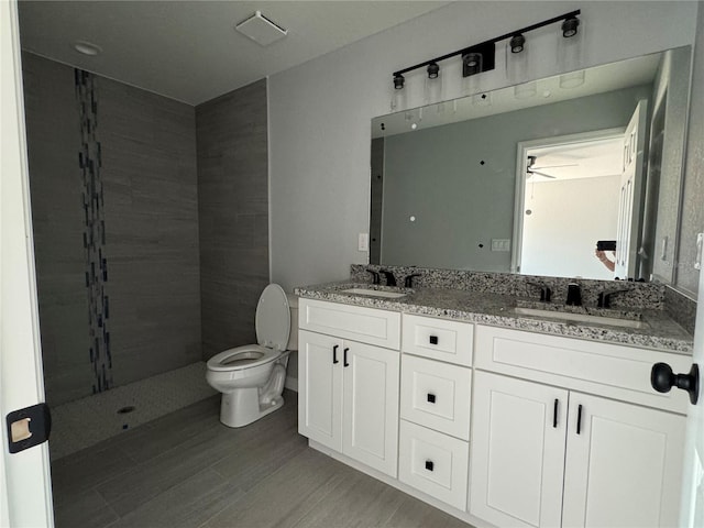 bathroom featuring toilet, wood finished floors, double vanity, ceiling fan, and a tile shower