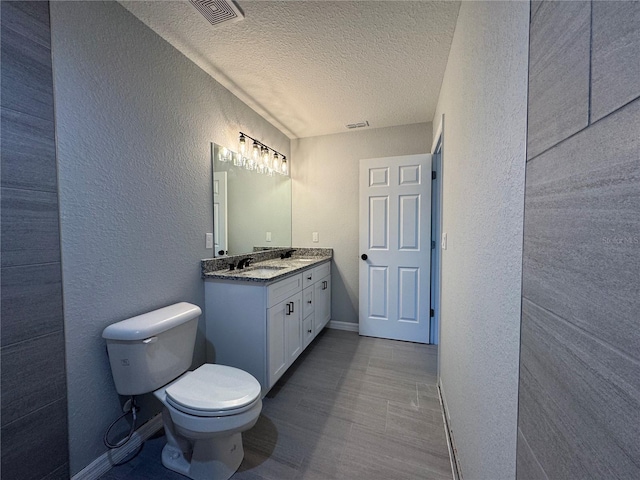 bathroom with a textured wall, a textured ceiling, toilet, baseboards, and double vanity