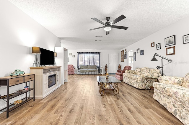 living room featuring a textured ceiling, a ceiling fan, light wood-type flooring, and a glass covered fireplace