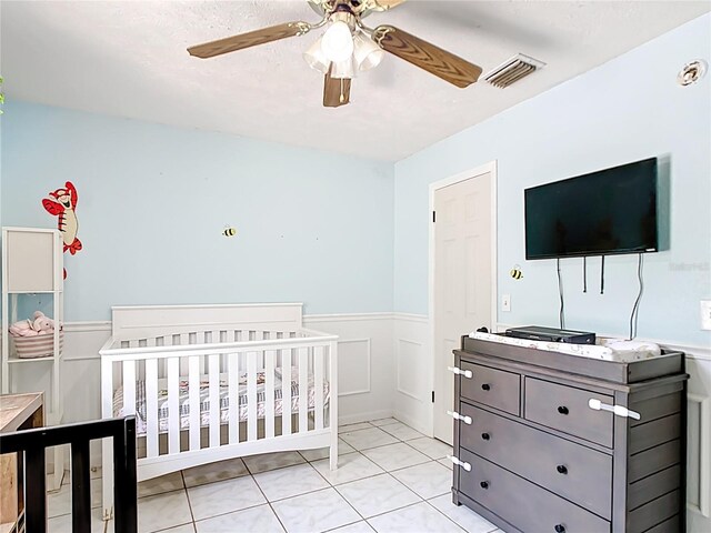 bedroom with a wainscoted wall, a crib, light tile patterned floors, a decorative wall, and a ceiling fan