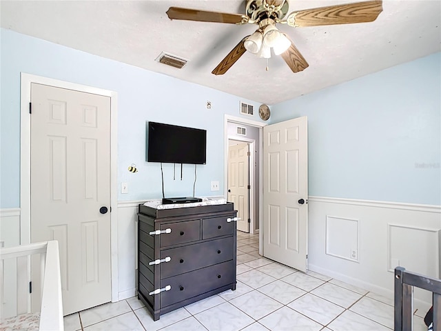 bedroom featuring wainscoting, light tile patterned flooring, ceiling fan, and a decorative wall