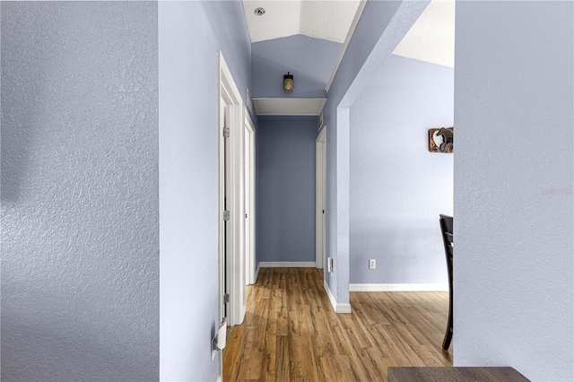 corridor with wood finished floors, a textured wall, baseboards, and vaulted ceiling