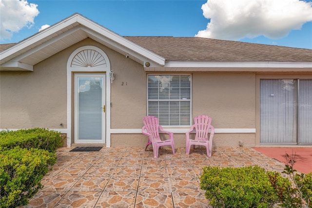 doorway to property with stucco siding, a patio area, and a shingled roof