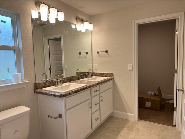 full bath featuring toilet, double vanity, a textured wall, and baseboards