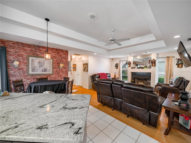 tiled living room with a tile fireplace, brick wall, ceiling fan, a textured ceiling, and a tray ceiling