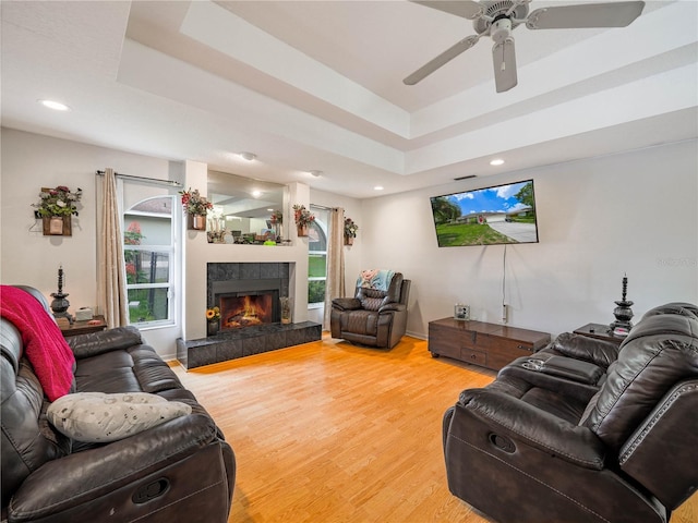 living room featuring ceiling fan, a tile fireplace, a raised ceiling, and light hardwood / wood-style flooring