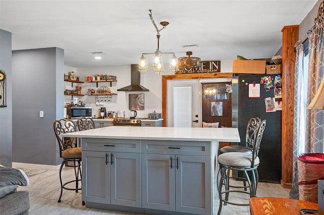 kitchen featuring gray cabinets, hanging light fixtures, light hardwood / wood-style floors, wall chimney range hood, and black fridge