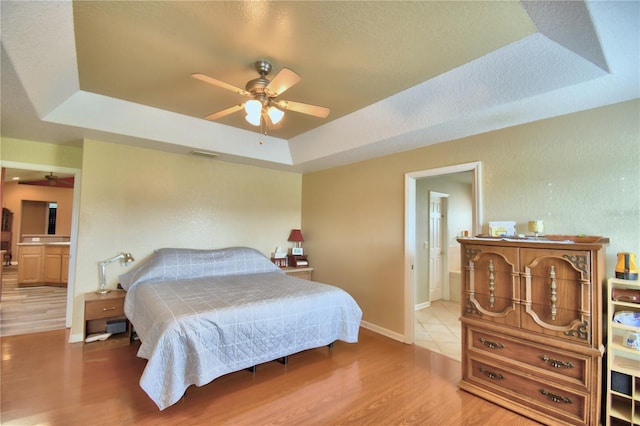 bedroom featuring ceiling fan, connected bathroom, a textured ceiling, light hardwood / wood-style flooring, and a tray ceiling