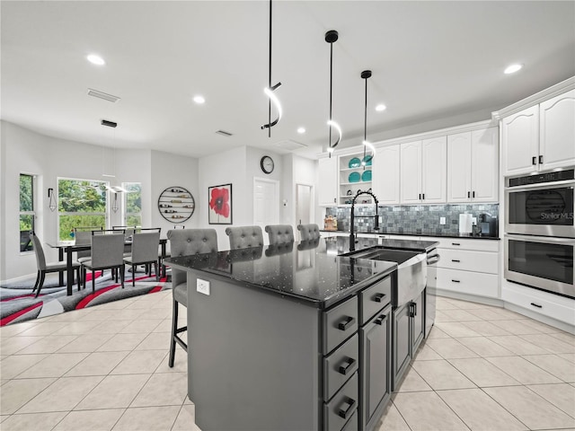 kitchen with an island with sink, hanging light fixtures, decorative backsplash, stainless steel double oven, and white cabinets