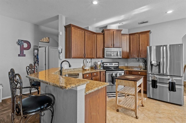 kitchen featuring a peninsula, a sink, light stone countertops, brown cabinetry, and appliances with stainless steel finishes