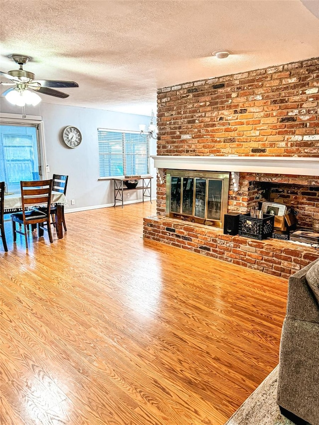 living area featuring wood finished floors, baseboards, a textured ceiling, a brick fireplace, and ceiling fan with notable chandelier